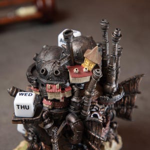 May include: A detailed, steampunk-inspired sculpture featuring a complex arrangement of pipes, gears, and miniature houses. The sculpture has a weathered, metallic finish with red and yellow accents. A white die displays the word "WED" and "THU".