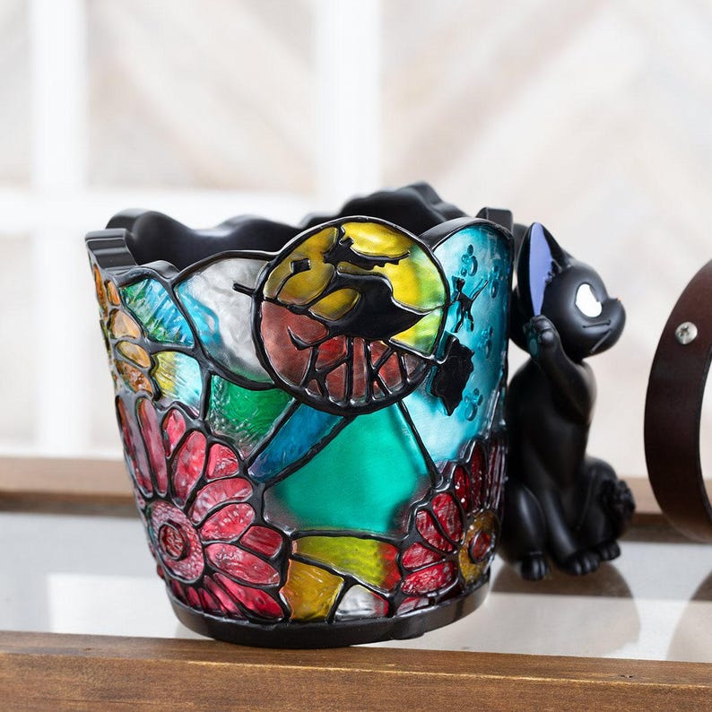 K&ouml;nnte beinhalten: Ein schwarzer Keramiktopf mit einem Buntglasdesign mit bunten Blumen, einer gelben Sonne und einer schwarzen Katze. Der Topf ist perfekt f&uuml;r die Pr&auml;sentation kleiner Pflanzen oder Sukkulenten.