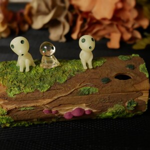 Pode incluir: Uma caixa decorativa de madeira com um topo verde musgo, apresentando tr&ecirc;s figuras Kodama. As figuras Kodama s&atilde;o brancas com olhos pretos. A caixa tem uma apar&ecirc;ncia natural de gr&atilde;o de madeira com detalhes em rosa.