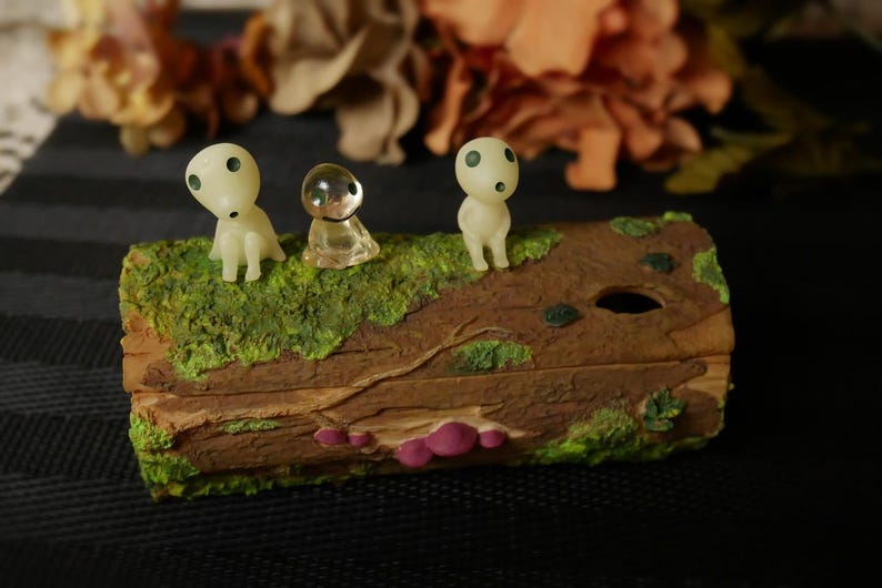 Pode incluir: Uma caixa decorativa de madeira com um topo verde musgo, apresentando tr&ecirc;s figuras Kodama. As figuras Kodama s&atilde;o brancas com olhos pretos. A caixa tem uma apar&ecirc;ncia natural de gr&atilde;o de madeira com detalhes em rosa.