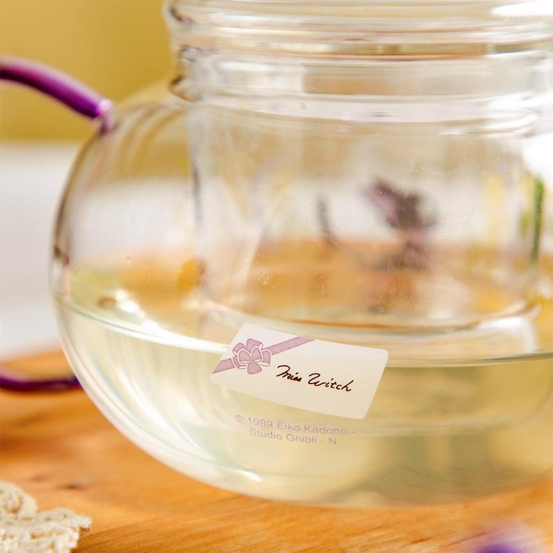 Puede incluir: Una tetera de cristal transparente con un asa morada y una etiqueta rectangular blanca con un gr&aacute;fico de lazo rosa y el texto Miss Witch. Debajo de la etiqueta, el cristal tiene grabado el texto 1989 Eiko Kadono - Studio Ghibli - N.