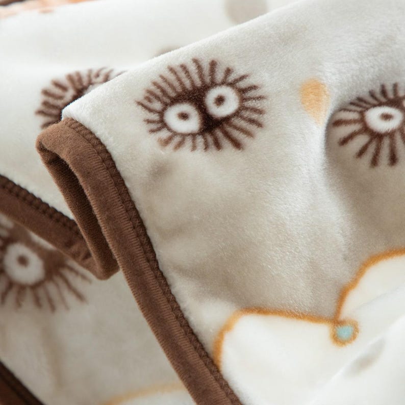 Peut inclure: Gros plan sur une couverture douce avec un motif r&eacute;p&eacute;t&eacute; de personnages de dessins anim&eacute;s bruns aux grands yeux. La couverture a un fond cr&egrave;me et une bordure marron.