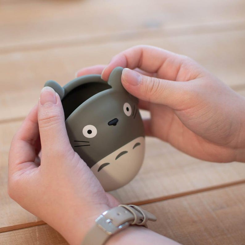 May include: A gray and white Totoro-shaped container with black details, including eyes, nose, and whiskers. The container has two small ears and a rounded shape. The container is being held by two hands.