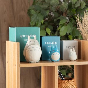 May include: A collection of Studio Ghibli Totoro figurines and boxes. The figurines are in various sizes and colors, including gray, blue, and white. The boxes are teal, blue, and gray, with Japanese text. The items are displayed on a wooden shelf.