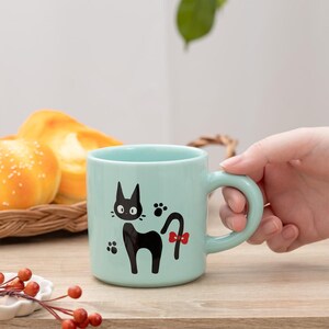 May include: A light mint green ceramic mug featuring a graphic illustration of a black cat with large eyes and a red bow on its tail. The design includes small black paw prints surrounding the cat. The mug has a smooth finish and a curved handle.