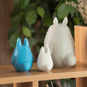 May include: Three ceramic figurines of the character Totoro from the animated film 'My Neighbor Totoro'. The figurines are in blue, white, and gray, and are arranged on a wooden shelf. The largest figurine is approximately 6 inches tall.