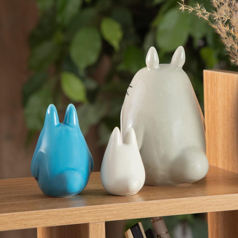 May include: Three ceramic figurines of the character Totoro from the animated film 'My Neighbor Totoro'. The figurines are in blue, white, and gray, and are arranged on a wooden shelf. The largest figurine is approximately 6 inches tall.