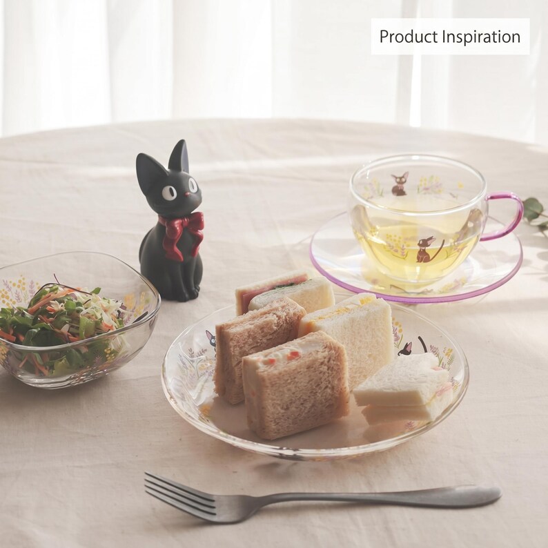 Op de afbeelding: Een zwart kattenbeeldje met een rode strik, een doorzichtig glazen theekopje en schoteltje met een paars handvat en rand, een glazen kom met salade, een glazen bord met vier kleine sandwiches en een metalen vork op de voorgrond.