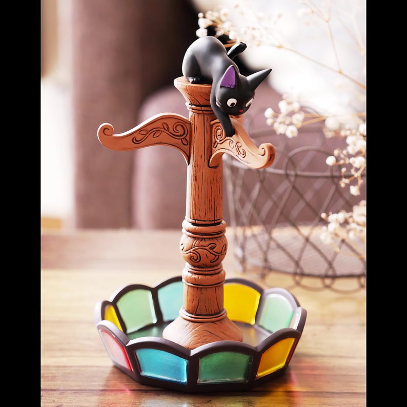May include: A decorative cat figurine perched on a brown lamp post-style holder with a colorful stained-glass base. The black cat has purple accents on its ears. The base features a variety of colors including red, green, blue, and yellow.