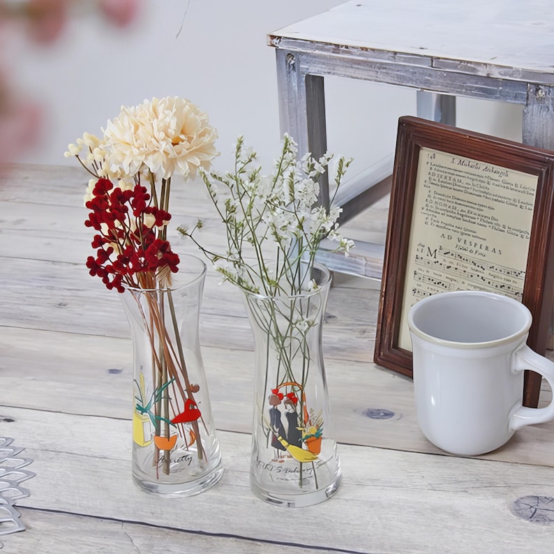 Puede incluir: Dos jarrones de cristal transparente con ilustraciones florales, cada uno con diferentes flores. Un jarr&oacute;n tiene flores rojas y crema, el otro flores blancas. Una impresi&oacute;n enmarcada y una taza blanca est&aacute;n en el fondo.