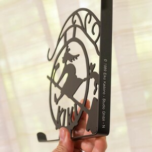 K&ouml;nnte beinhalten: Schwarzer Metall-Wandhaken mit einer Silhouette einer Hexe auf einem Besen in einem kreisf&ouml;rmigen Rahmen. Der Haken hat eine gebogene Unterseite und eine rechteckige R&uuml;ckseite. Der Text auf der Seite lautet "&copy; 1989 Eiko Kadono - Studio Ghibli - N."