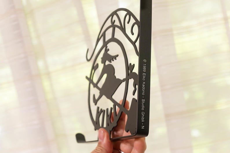 K&ouml;nnte beinhalten: Schwarzer Metall-Wandhaken mit einer Silhouette einer Hexe auf einem Besen in einem kreisf&ouml;rmigen Rahmen. Der Haken hat eine gebogene Unterseite und eine rechteckige R&uuml;ckseite. Der Text auf der Seite lautet "&copy; 1989 Eiko Kadono - Studio Ghibli - N."
