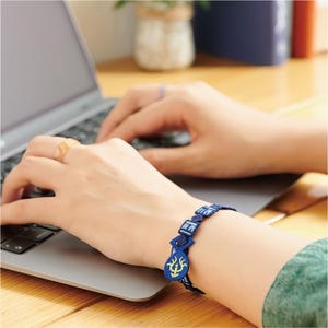 K&ouml;nnte beinhalten: Ein blau besticktes Armband mit geometrischen und floralen Designs. Das Armband wird am Handgelenk getragen, mit einem Laptop im Hintergrund. Das Armband hat eine dunkelblaue Basis mit hellblauen und gelben Akzenten.