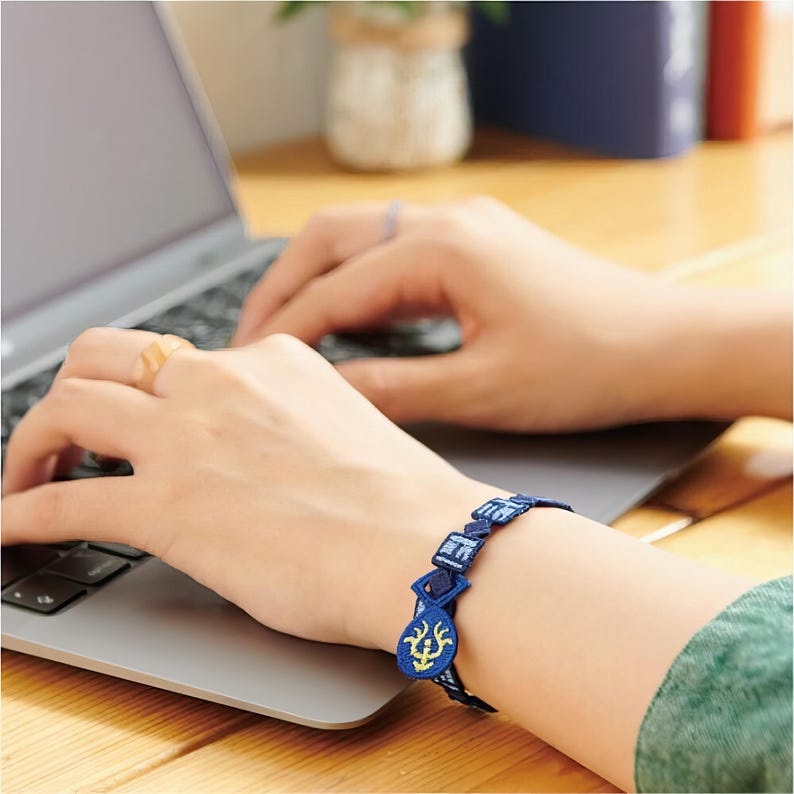 K&ouml;nnte beinhalten: Ein blau besticktes Armband mit geometrischen und floralen Designs. Das Armband wird am Handgelenk getragen, mit einem Laptop im Hintergrund. Das Armband hat eine dunkelblaue Basis mit hellblauen und gelben Akzenten.