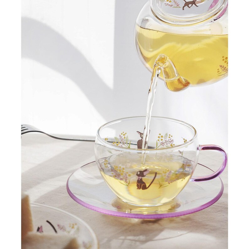 Puede incluir: Una tetera de cristal transparente vierte t&eacute; amarillo claro en una taza y platillo a juego. El conjunto presenta un delicado estampado de un peque&ntilde;o gato negro rodeado de ramitas de flores amarillas y moradas. La taza tiene un asa morada.