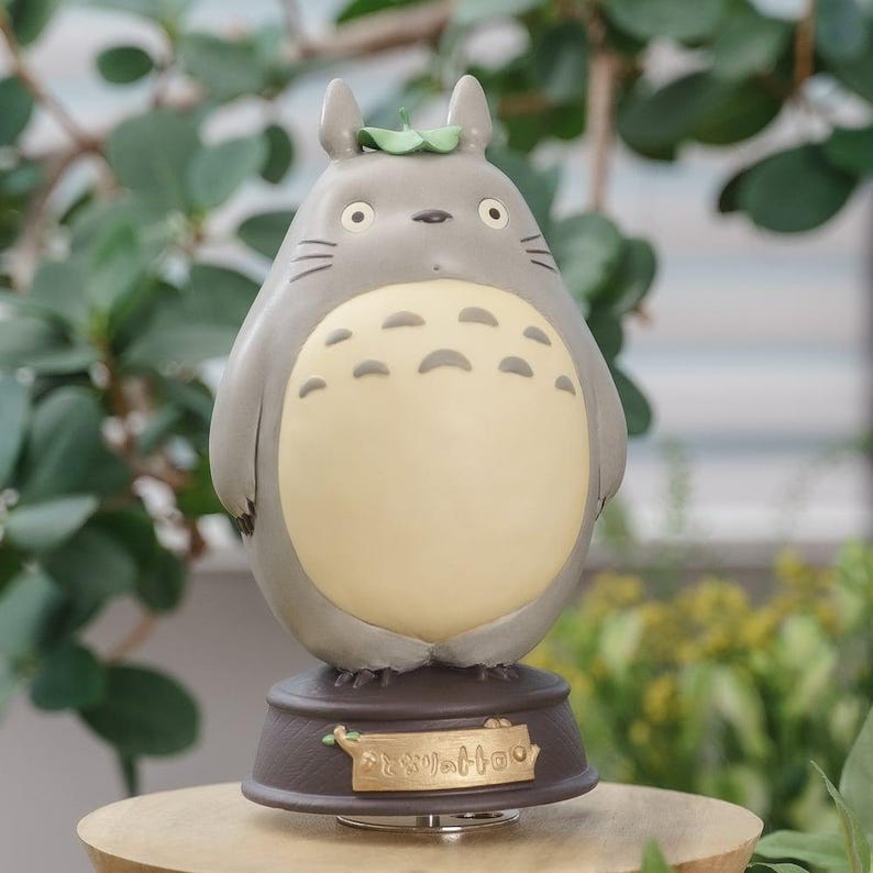 May include: A gray Totoro figurine with a cream-colored belly, small ears, and whiskers. The figurine has a leaf on its head and sits on a brown base with Japanese text. The background features green foliage.
