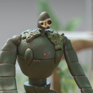 Peut inclure: Figurine d&eacute;taill&eacute;e d'un personnage robot, principalement vert olive avec des d&eacute;tails dor&eacute;s. Le robot a un corps arrondi, des membres articul&eacute;s et une t&ecirc;te avec un design unique en forme d'&oelig;il. Le haut du corps du robot est orn&eacute; d'une texture semblable &agrave; de la mousse.