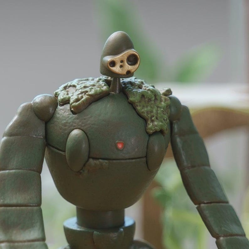 Peut inclure: Figurine d&eacute;taill&eacute;e d'un personnage robot, principalement vert olive avec des d&eacute;tails dor&eacute;s. Le robot a un corps arrondi, des membres articul&eacute;s et une t&ecirc;te avec un design unique en forme d'&oelig;il. Le haut du corps du robot est orn&eacute; d'une texture semblable &agrave; de la mousse.