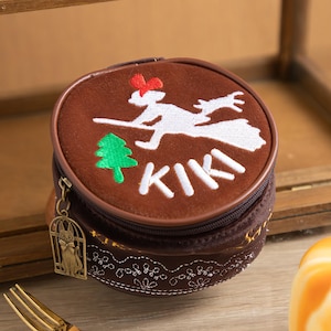 May include: A round, brown coin purse with a zipper closure. The purse features an embroidered design of a witch on a broom, a green tree, and the word "KIKI" in white. A small charm of a cat in a cage hangs from the zipper.