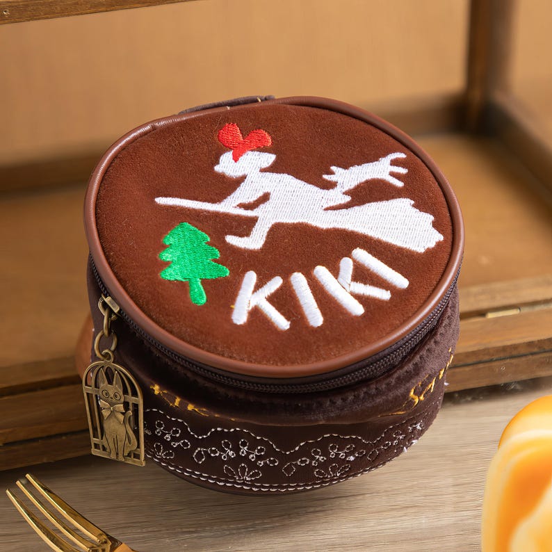 May include: A round, brown coin purse with a zipper closure. The purse features an embroidered design of a witch on a broom, a green tree, and the word "KIKI" in white. A small charm of a cat in a cage hangs from the zipper.
