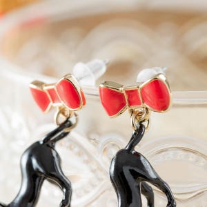 May include: A pair of gold-toned earrings featuring red bow accents and black cat silhouettes. The earrings have post-style backs and are set against a soft, blurred background.