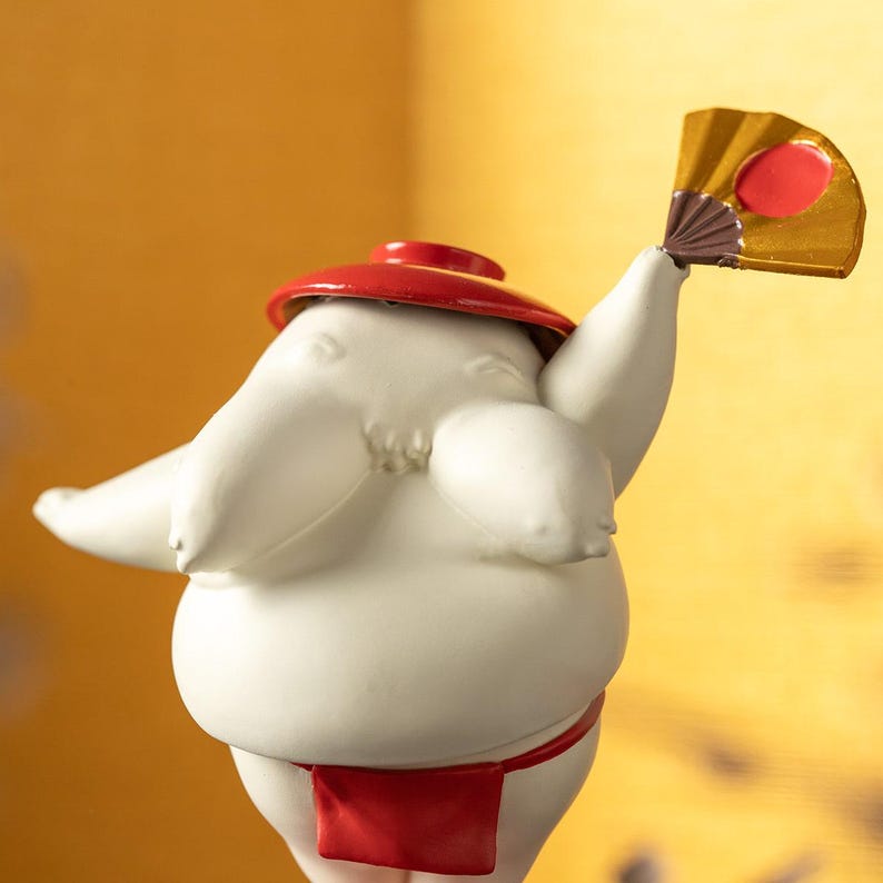 Peut inclure: Figurine en c&eacute;ramique d'un lutteur de sumo, vue de dos, portant un mawashi et un chapeau rouges. Le lutteur tient un &eacute;ventail dor&eacute; avec un cercle rouge. La figurine est blanche avec des accents rouges et a une finition lisse.