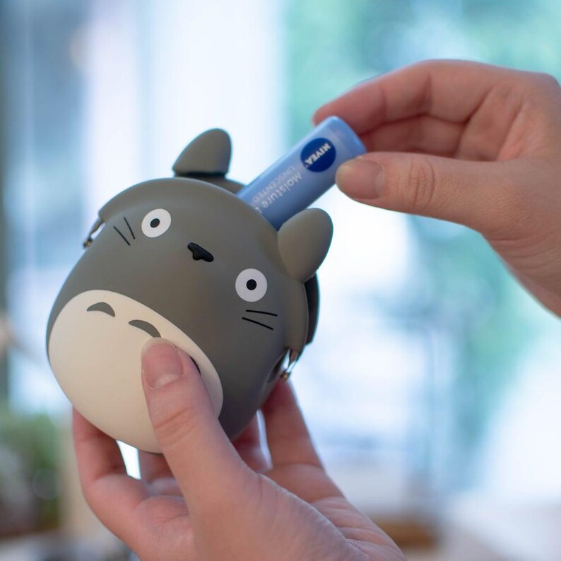 May include: A gray Totoro-shaped coin purse with a white belly and black details. A blue Nivea lip balm is being placed inside the purse. The purse has a zipper closure and small silver metal accents.