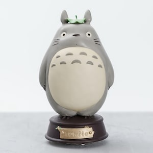May include: A gray Totoro figurine with a cream-colored belly and a green leaf on its head. The figurine sits on a brown base with Japanese text. The figure has black whiskers and large, round eyes.