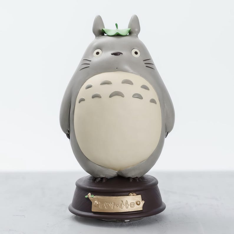 May include: A gray Totoro figurine with a cream-colored belly and a green leaf on its head. The figurine sits on a brown base with Japanese text. The figure has black whiskers and large, round eyes.