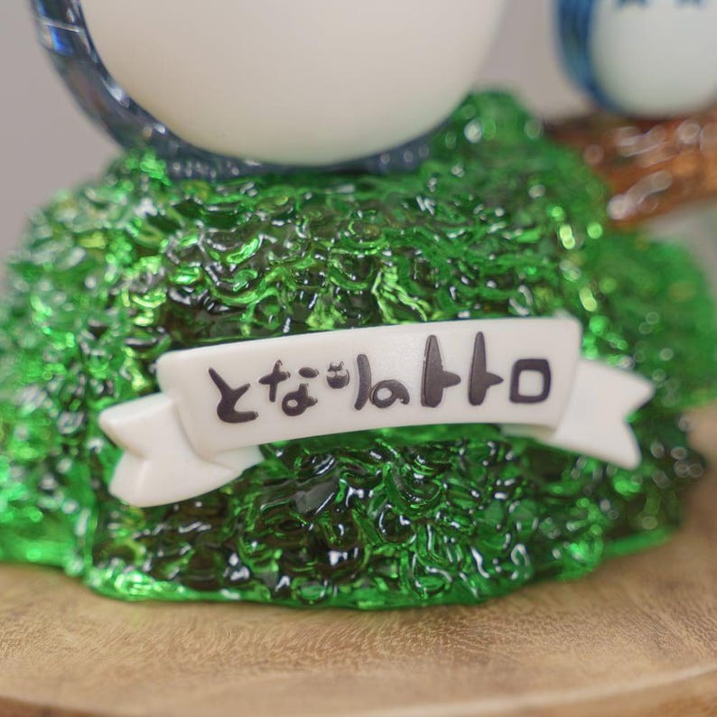 Op de afbeelding: Een close-up van een beeldje met een banner met de Japanse tekst "となりのトトロ" in het zwart. De basis is een getextureerd, doorschijnend groen, dat op bladeren lijkt. De banner is wit.