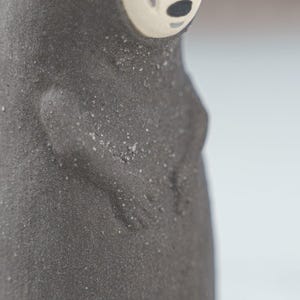 Puede incluir: Una figura de cer&aacute;mica gris oscuro con una superficie texturizada y granulada. El objeto presenta un rostro liso de color claro con nariz y boca negras. El torso muestra brazos esculpidos que descansan contra el cuerpo, con un acabado granulado.