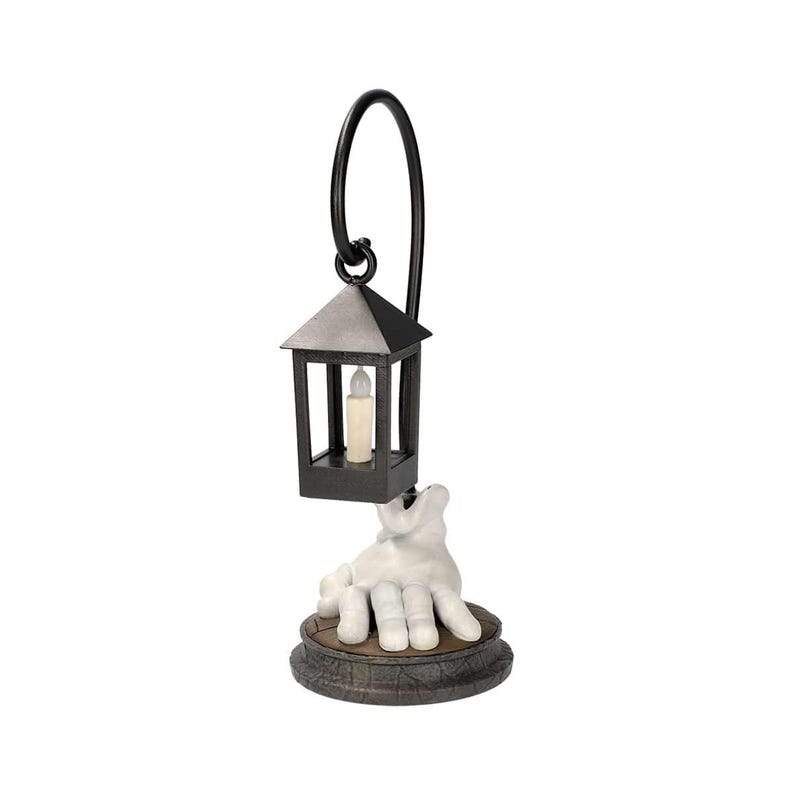 May include: A decorative lantern with a black frame and a faux candle inside. The lantern is suspended from a curved black metal handle and rests above a white hand sculpture. The hand is emerging from a round, textured base.