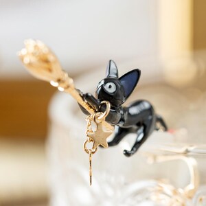 May include: A black cat figurine with blue-tipped ears, holding a gold broom. A gold star charm hangs from the cat's collar. The cat is posed as if flying, with a whimsical and magical theme.