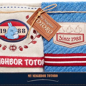 Puede incluir: Dos toallas de felpa con el personaje animado Totoro. Una es beige con un gr&aacute;fico de Totoro azul y detalles en rojo. La otra es azul con una raya roja y blanca. Ambas tienen el texto "My Neighbor Totoro."