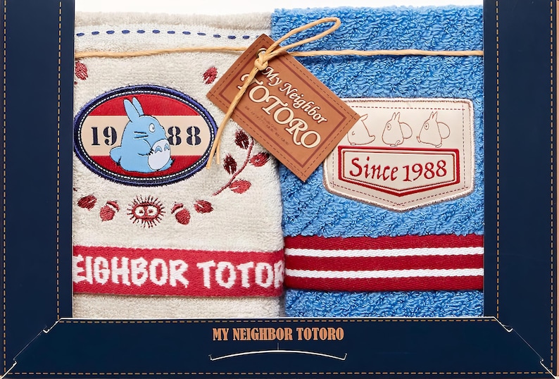 Puede incluir: Dos toallas de felpa con el personaje animado Totoro. Una es beige con un gr&aacute;fico de Totoro azul y detalles en rojo. La otra es azul con una raya roja y blanca. Ambas tienen el texto "My Neighbor Totoro."