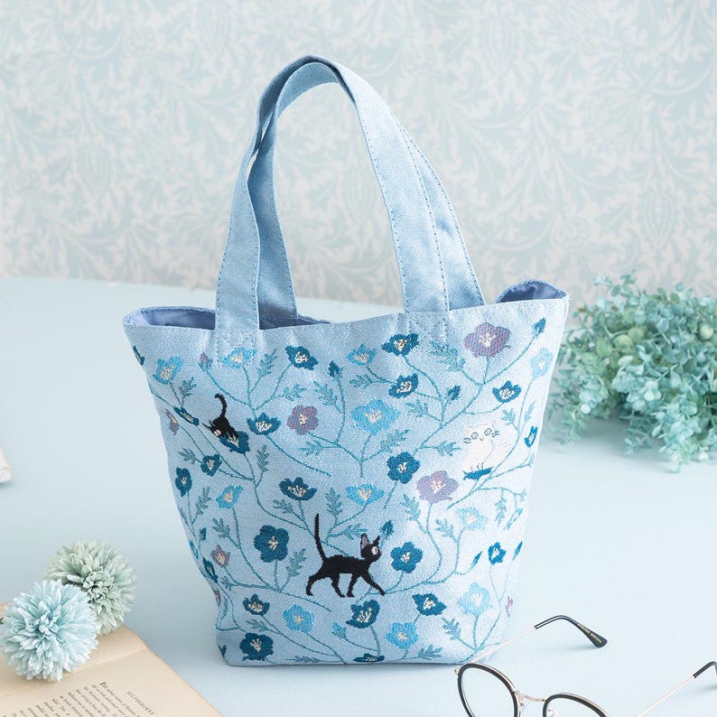May include: A light blue tote bag with embroidered black cats and blue flowers. The bag has two fabric handles.