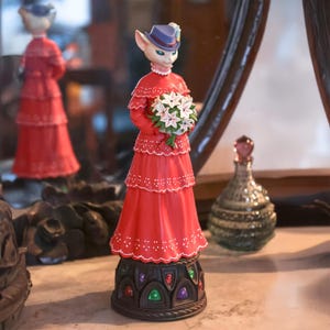 Puede incluir: Una figura caprichosa de un personaje felino con un vestido rojo y un sombrero azul, sosteniendo un ramo de flores blancas. La figura tiene una base decorativa con detalles coloridos. Un espejo refleja la figura.