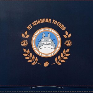 Puede incluir: Una caja azul marino con las palabras "MY NEIGHBOR TOTORO" y un emblema circular con Totoro. El emblema est&aacute; rodeado por una corona de hojas y bellotas. La caja tiene detalles en las esquinas de color beige.