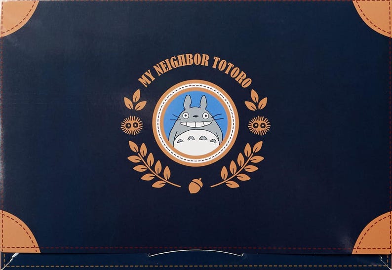 Puede incluir: Una caja azul marino con las palabras "MY NEIGHBOR TOTORO" y un emblema circular con Totoro. El emblema est&aacute; rodeado por una corona de hojas y bellotas. La caja tiene detalles en las esquinas de color beige.