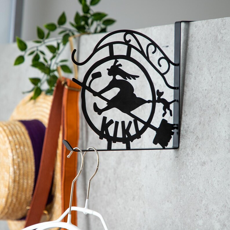 K&ouml;nnte beinhalten: Schwarzer Metall-T&uuml;rh&auml;nger mit einer Silhouette einer Hexe auf einem Besen und einer Katze, sowie dem Wort "KIKI". Der H&auml;nger hat ein dekoratives Scroll-Design. Geeignet f&uuml;r Kinderzimmer oder Halloween-Dekoration.