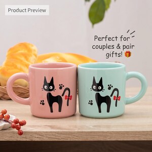 May include: Two ceramic mugs, one pastel pink and one light blue, each featuring a black cat illustration with a red bow on its tail and small paw prints. The mugs have rounded handles and a smooth finish, displayed side by side on a surface.