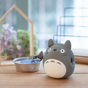 May include: A gray Totoro-shaped coin purse with a white belly and black details. The coin purse has two small ears and a slot for coins. It is sitting on a wooden surface.