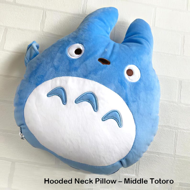 May include: A blue plush hooded neck pillow shaped like a cartoon character, featuring white accents on the belly and eyes. The pillow has a rounded shape with two upright ears and a small brown nose. The text "Hooded Neck Pillow - Middle Totoro" is at the bottom.