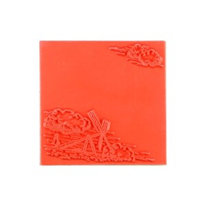 May include: A square red rubber stamp featuring a raised design of a windmill in the bottom left corner and a small cloud formation in the top right corner. The surface is flat and orange-red, with the embossed imagery creating a textured relief.