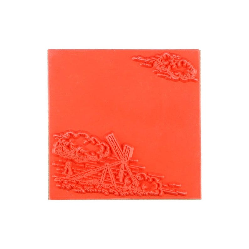 May include: A square red rubber stamp featuring a raised design of a windmill in the bottom left corner and a small cloud formation in the top right corner. The surface is flat and orange-red, with the embossed imagery creating a textured relief.
