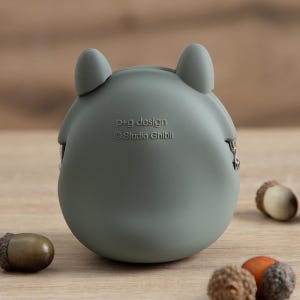 May include: A gray, rounded coin purse with two small, upright ear-like features. The purse has the text "p+g design" and "&copy; Studio Ghibli" printed on it. Several acorns are scattered around the purse on a wooden surface.