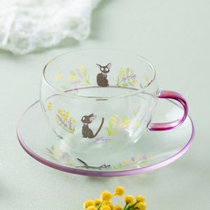 Op de afbeelding: Een set van een doorzichtige glazen theekop en schotel met een delicaat bloemmotief van gele en groene takjes. Het ontwerp bevat kleine zwarte katillustraties met roze accenten. Het oortje van de kop en de rand zijn paars getint.