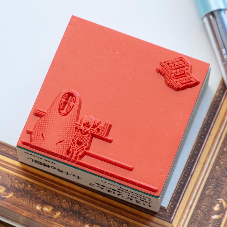 May include: A square rubber stamp with a red surface featuring embossed designs of a hooded figure and a small character. The side of the wooden base displays black text including "Studio Ghibli" and Japanese characters, with "MADE IN CHINA" printed.