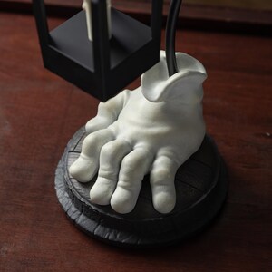 May include: A unique, decorative lamp base featuring a sculpted, white hand emerging from a dark, round base. The hand appears to be holding a black lamp fixture. The base has a textured, wooden appearance, and the overall design is artistic and unusual.