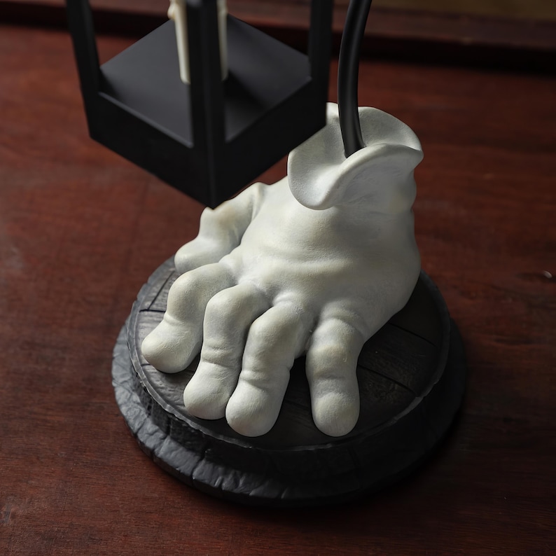 May include: A unique, decorative lamp base featuring a sculpted, white hand emerging from a dark, round base. The hand appears to be holding a black lamp fixture. The base has a textured, wooden appearance, and the overall design is artistic and unusual.